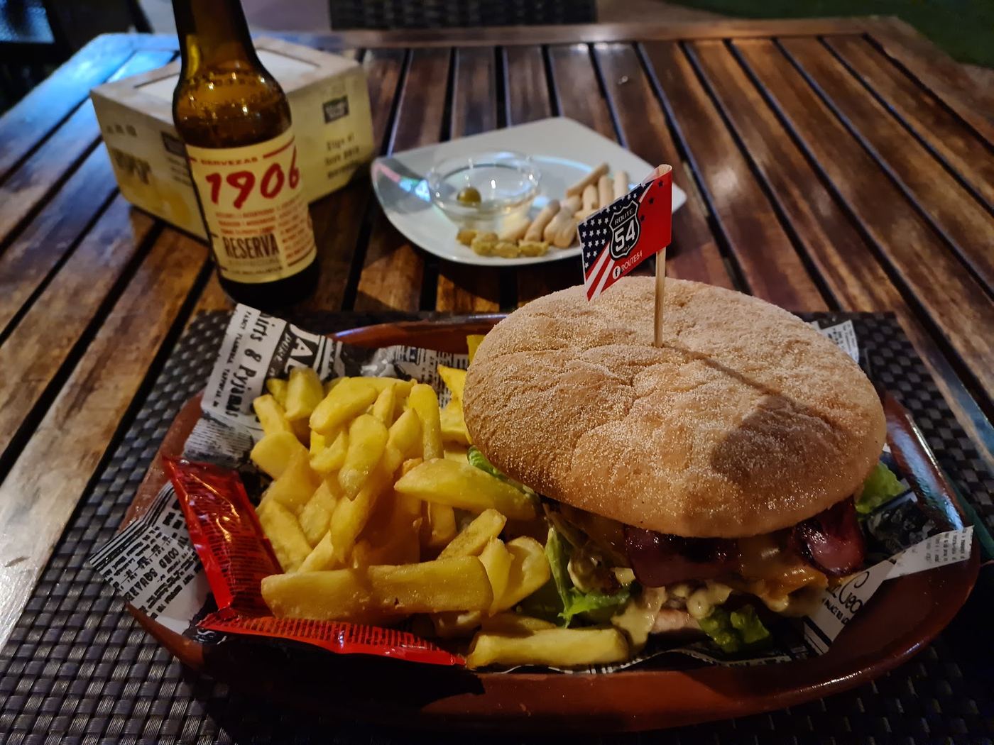 Hamburguesa clásica con queso, lechuga y tomate, servida con patatas fritas y una cerveza 1906 Reserva.